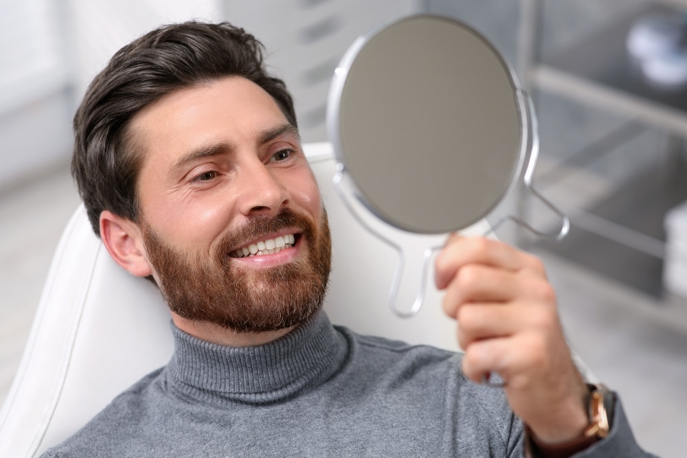 Happy man checking teeth in mirror at dentist